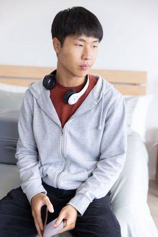 Adult asian man sitting on bed in hotel wearing gray hoodie and headphones holding phone