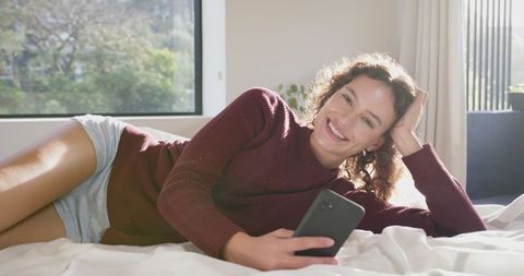 Smiling Woman Relaxing on Bed with Smartphone in Sunny Bedroom