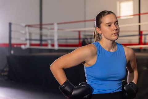 Determined female boxer standing hands on hips wearing blue tank top and black gloves beside boxing