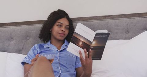 African american woman relaxing in satin pajamas reading book in bed with tea