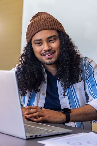 Asian young man working remotely on laptop at desk wearing brown beanie and striped shirt