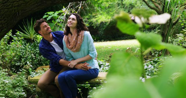 A young Caucasian couple enjoys a moment together on a bench in a lush green garden, with copy space. Their relaxed postures and happy expressions suggest a romantic connection in a serene outdoor setting.