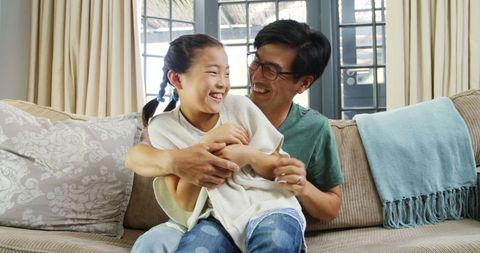 Father tickles daughter on sofa enjoying familial bond