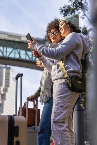 Two women traveling city checking smartphone with luggage near urban skywalk wearing hoodies