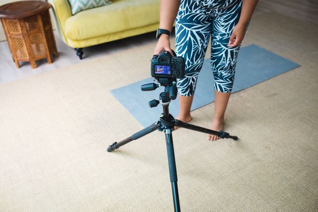 African American woman adjusting digital camera on tripod, preparing to record a home fitness video. Ideal for content related to fitness, healthy lifestyle, home workouts, blogging, and social media influencing.