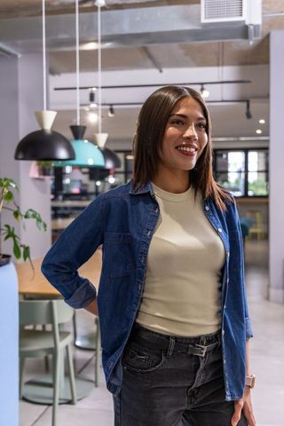 Smiling asian woman standing in modern coworking lounge wearing denim shirt and wristwatch