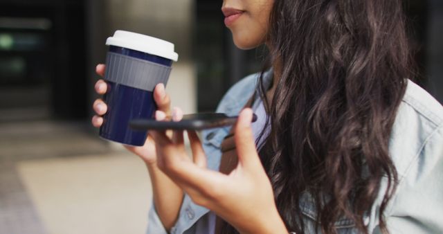 A woman is standing in an urban area, holding a takeaway coffee cup and smartphone. She is speaking into or using her phone while enjoying her coffee. Ideal for content on modern lifestyle, technology, communication, on-the-go living, and casual urban scenes.