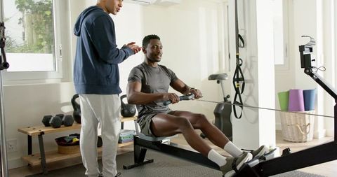 African american man rowing on indoor rower with personal trainer coaching at home gym