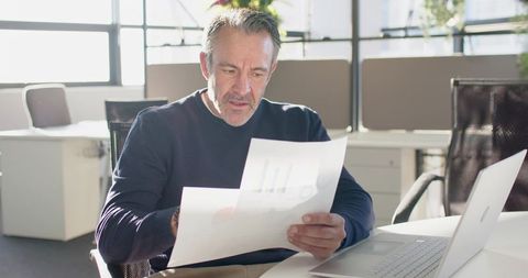 Mature Professional Reviewing Financial Charts at Modern Open-Plan Desk with Laptop