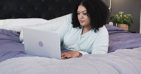 Mid-30s woman reclining on bed using laptop in cozy pastel pajamas at home