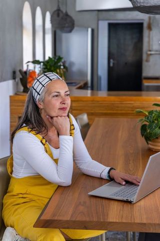 Thoughtful mature woman working at laptop in modern rustic kitchen with yellow overalls