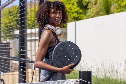 Young woman carrying padel racket at outdoor court smiling with headphones and backpack