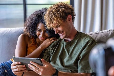 Couple cuddling on sunlit couch while watching tablet, sharing laugh and cozy warm moment