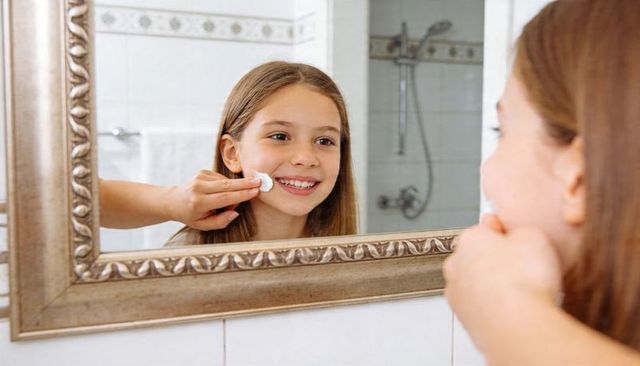 Smiling girl applying gentle skincare with cotton pad at ornate bathroom mirror reflection