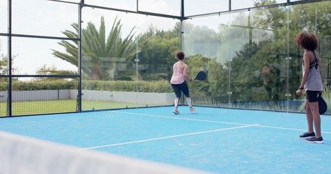 African american women playing padel on outdoor glass court with blue turf and paddles