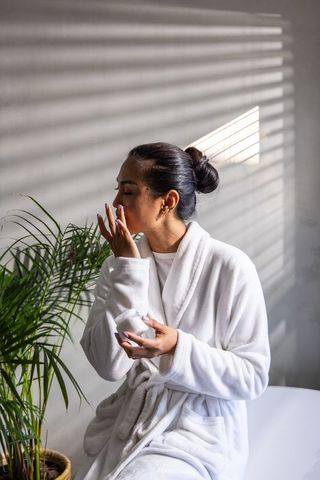 Asian woman in white bathrobe applying moisturizer on face in sunlit minimalist bedroom
