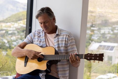 Mature man playing acoustic guitar on sunlit window seat overlooking suburban hillside