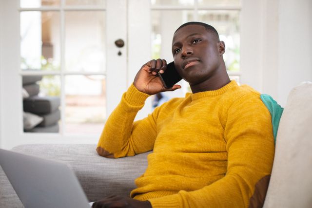 Young African American man multitasking by talking on smartphone and using laptop while sitting on a couch at home. Ideal for illustrating remote work, modern communication, and casual home lifestyle. Suitable for articles, blogs, and advertisements related to technology, work-from-home setups, and everyday life.