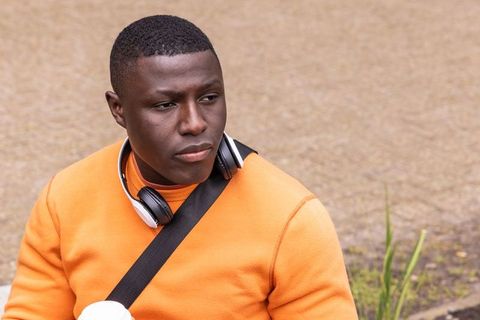 African american man sitting on pavement wearing orange sweater headphones holding coffee