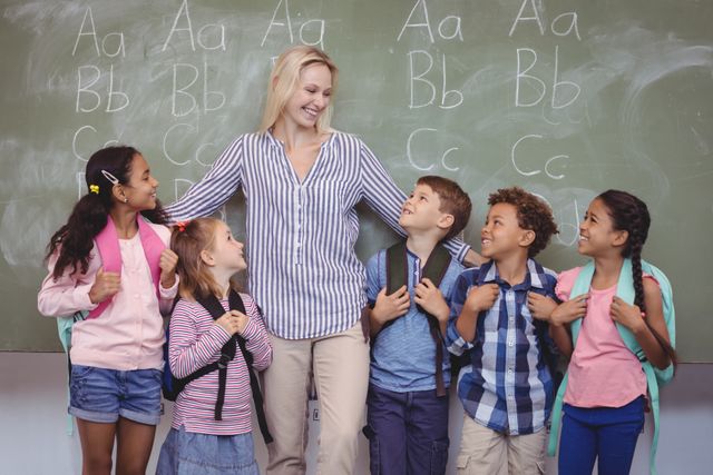 Teacher standing with diverse group of schoolchildren in classroom, all smiling and wearing backpacks. Chalkboard in background with alphabet letters. Ideal for educational materials, school promotions, and diversity in education themes.