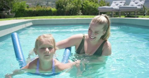 Smiling parent teaching child to swim with blue noodle in sunny backyard pool