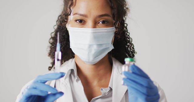 Image of portrait of biracial female doctor with face mask holding vaccine vial on white background. global medicine and healthcare concept.