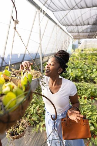 African american woman inspecting hanging planter in greenhouse nursery holding leather bag