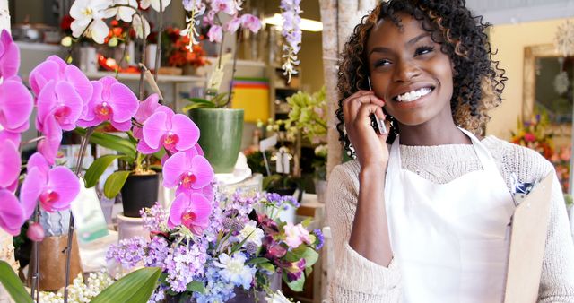 A young African American woman smiles brightly as she talks on the phone in a vibrant flower shop, with copy space. Her joyful expression and the colorful floral background suggest a pleasant conversation amidst a cheerful work environment.