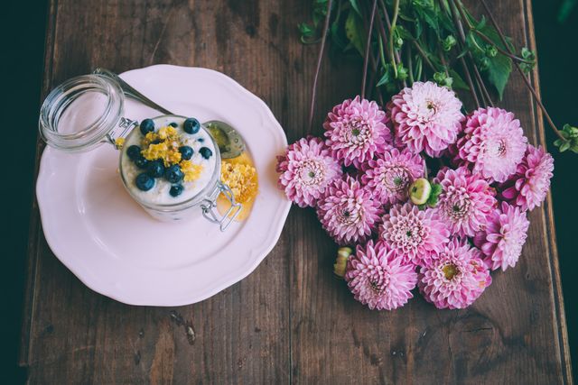 Glass jar filled with layers of creamy yogurt, fresh blueberries, and edible flowers on white plate. Bright pink zinnias laying on wooden surface next to jar. Ideal for food blogs, health websites, home decor ideas, and breakfast inspiration sections.
