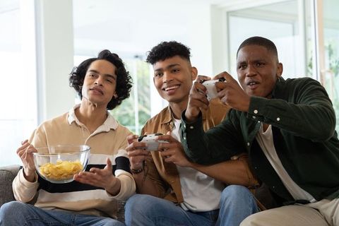 Three diverse young men playing video games on couch sharing bowl of chips in bright living room