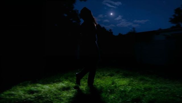 Moonlit backyard silhouette of hooded woman walking on grass at night casting long shadow
