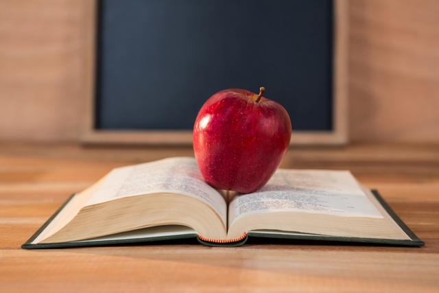 Red apple resting on an open book on a wooden table with a blackboard in the background. Ideal for educational themes, back-to-school promotions, healthy eating in schools, and academic materials.