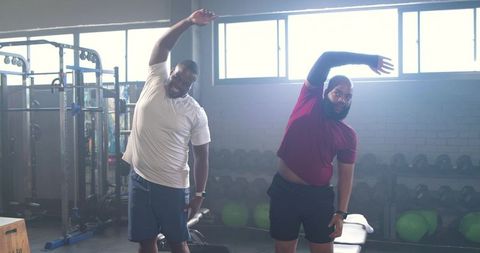 African male workout partners doing lateral stretches together in modern gym for mobility