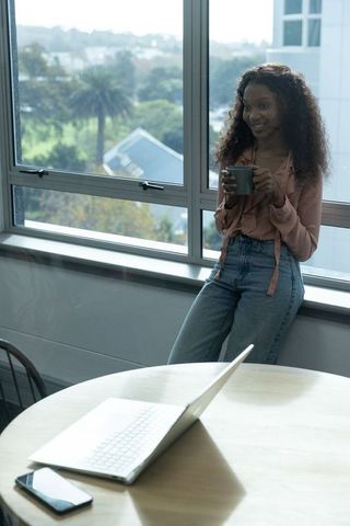 African-american woman holding mug, leaning on windowsill with laptop, relaxed work-from-home
