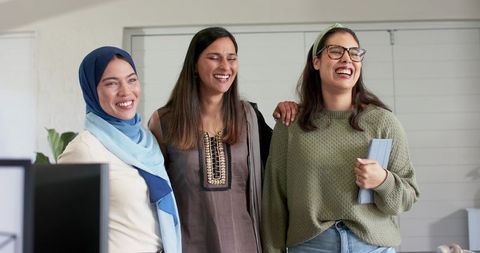 Multicultural women laughing and connecting in modern office holding tablet and computer