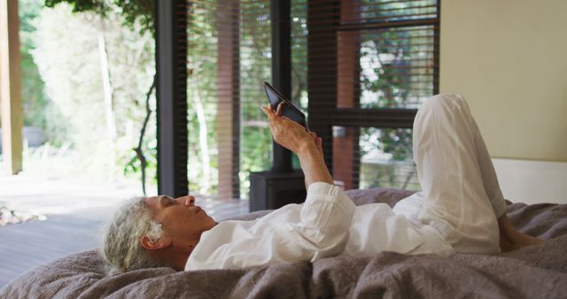 Senior woman lying in bed, using a tablet device comfortably. Natural light and serene outdoor backdrop coming through large windows, suggesting a peaceful morning or afternoon. Perfect for illustrating themes related to technology use among older adults, relaxation, home life, and leisure time. Suitable for websites and publications focused on seniors, home comfort, caregiving, lifestyle, and digital technology for the elderly.