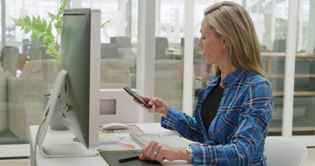 Caucasian woman reviews data on her phone in an office. She's focused on multitasking with technology at her modern workplace.