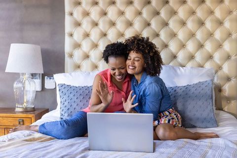 Smiling diverse female couple waving at laptop while relaxing on bed in cozy bedroom