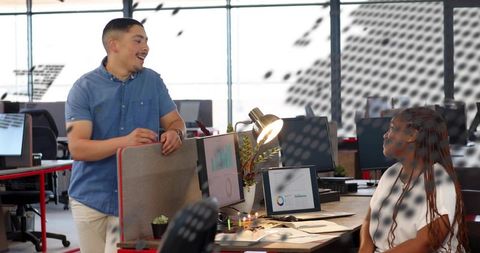 Smiling coworker leaning over partition collaborating with colleague at open-plan desk