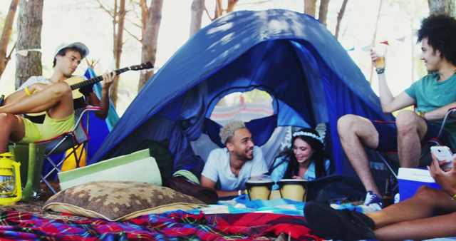 A diverse group of young adults enjoys a relaxed camping atmosphere, with one playing the guitar and others engaged in conversation. Their casual outdoor setting conveys a sense of friendship and leisure during a camping trip.