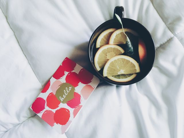 Stock photo showing a cozy start of the day with a warm cup of lemon tea on white bedding. Nearby, a colorful greeting card with the word 'hello,' enhancing the friendly and inviting mood. This photo captures a calm and peaceful morning routine ideal for themes related to relaxation, home comforts, self-care, morning rituals, or casual note writing.