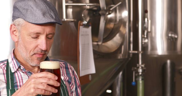 Male brewer savoring taste of freshly brewed beer at brewery. Standing next to large stainless steel brewing tank with tools for measuring quality. Great for use in articles or advertisements related to craft beer, beer brewing process, brewery tours, or quality control in beer production.