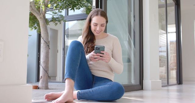 Young woman sitting on the floor near large windows, smiling and using her smartphone. She appears relaxed and comfortable in a modern interior space, which suggests a casual, at-home environment. Perfect for illustrating themes of connectivity, leisure, and modern living. Could be used in articles, advertisements or websites related to technology, home leisure activities, or millennial lifestyles.