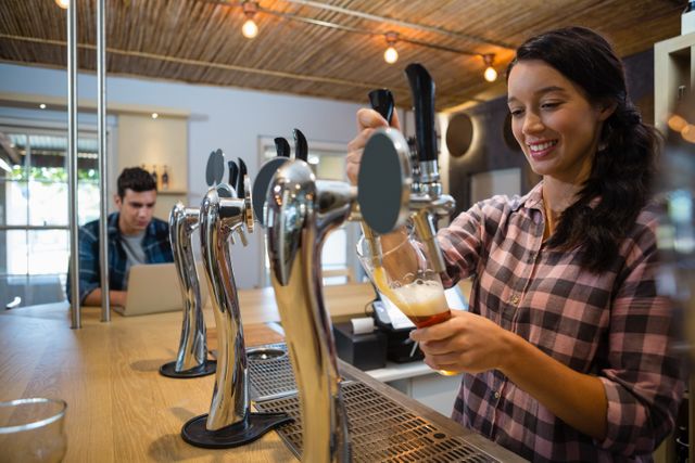 Young barmaid pouring beer from tap into glass in restaurant. Ideal for use in articles about hospitality industry, bar and restaurant services, customer service training, and promotional materials for pubs and bars.
