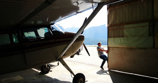 This image depicts a pilot opening a hangar door with an airplane inside on a sunny day. The airplane is partly visible with its nose pointing out. In the background, there are mountains and a clear blue sky, suggesting a scenic and pleasant environment typical of small airfields. This visual is excellent for illustrating themes related to aviation, travel, airfield operations, airport maintenance, and outdoor activities. It can be used for travel websites, aviation publications, brochures, advertisements, and any materials needing a vibrant outdoor aviation scene.