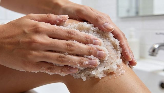 Hands applying coarse salt body scrub on knee closeup exfoliating skin care routine