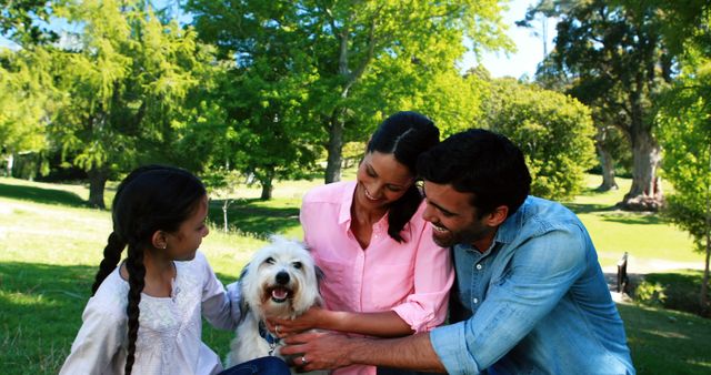 Family enjoying a beautiful day together in a green park setting surrounded by nature and trees. Parents and daughter nurturing their pet dog, all smiling pleasurably. Ideal for use in family-oriented advertisements, pet care promotions, outdoor leisure marketing, and wholesome lifestyle campaigns.