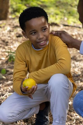 African american boy kneeling holding lemon while caregiver resting hand on shoulder in park