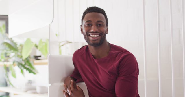 Smiling young man sitting indoors with natural light. Perfect for use in lifestyle, home living, advertising, and personal development materials conveying a positive and friendly atmosphere.