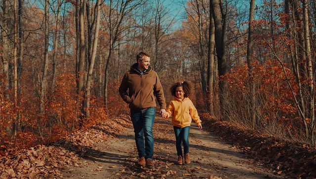 Father and daughter walking hand-in-hand on sunlit autumn forest trail, fall family outing
